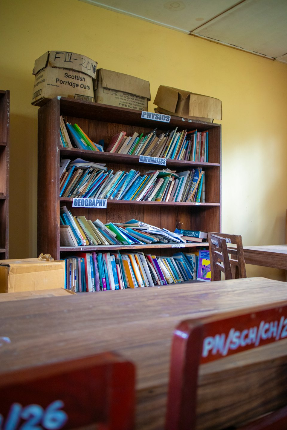 Ein überladenes Holzregal voller Bücher und Kisten in einem Klassenzimmer.